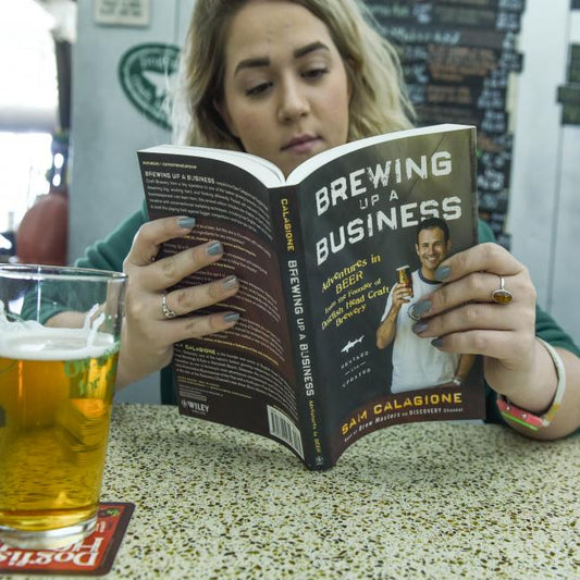 A woman sits at a table reading Brewing Up A Business by J Wiley and Sons Publishing, with a pint of craft beer in a glass on a coaster in front of her.