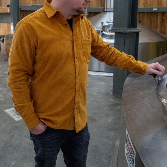 A person wearing the Brewery Branding, Inc. Camel Cord Flannel Button Down and jeans stands by a large metal brewing tank, holding a valve handle in an industrial space with concrete floors and wooden walls.