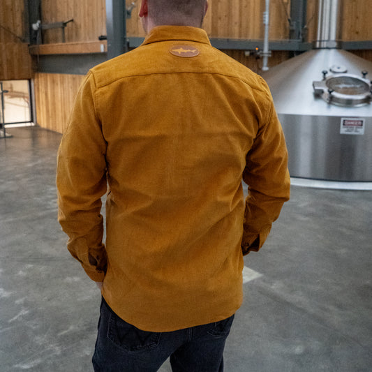 A person wearing the Camel Cord Flannel Button Down by Brewery Branding, Inc. stands with their back to the camera in an industrial space with brewing equipment and wooden walls.