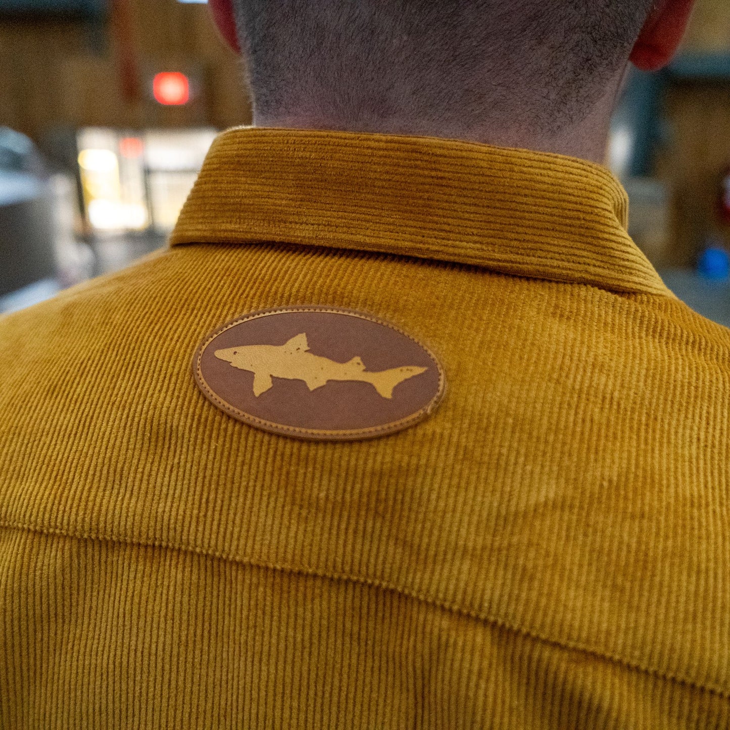 A person wears the Camel Cord Flannel Button Down by Brewery Branding, Inc., featuring camel-toned corduroy and a round brown shark silhouette patch on the upper back. The image highlights both the patch detail and textured fabric.