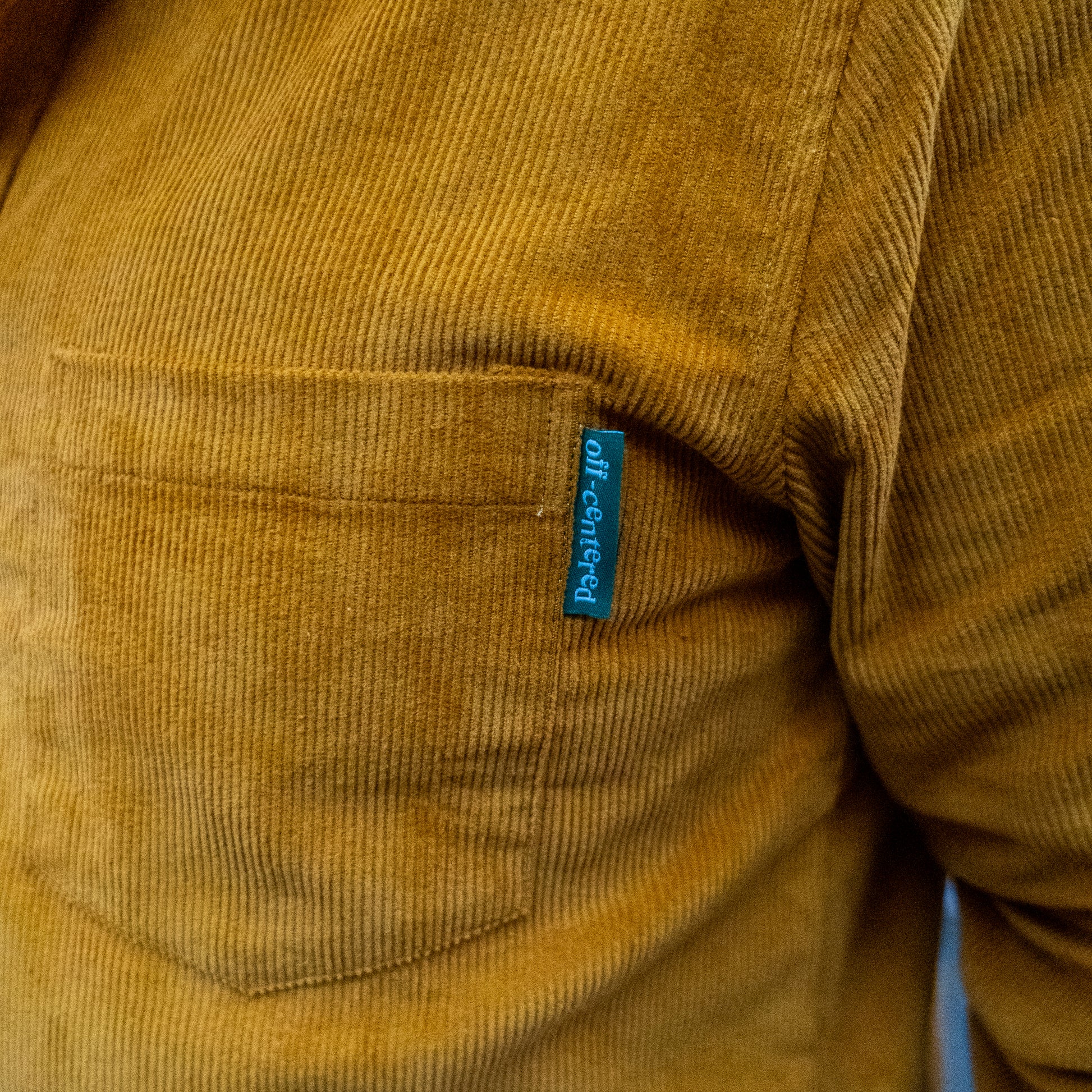 A close-up of the Camel Cord Flannel Button Down by Brewery Branding, Inc. shows its mustard yellow corduroy pocket with camel tones and a small blue off-centered tag.