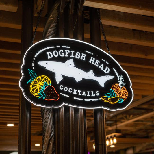 A vibrant Merchtank Canned Cocktails LED Sign features a dogfish shark silhouette and colorful fruit illustrations, including strawberry, lemon, and orange, celebrating the brand’s cocktails from a wooden-beamed ceiling.