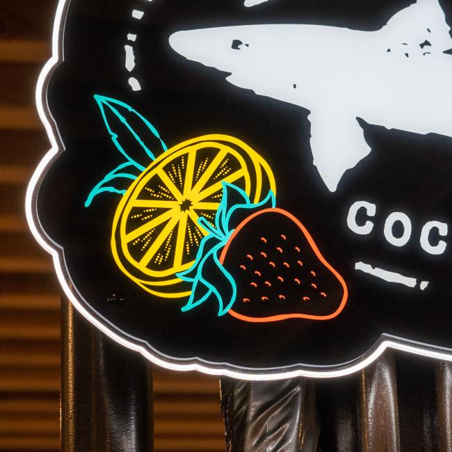 The Canned Cocktails LED Sign by Merchtank showcases a sliced lemon with leaves and a whole strawberry in vivid yellow, green, blue, and red outlines on black—a lively tribute to canned cocktails.