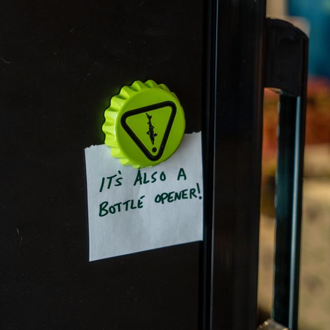The Caution Cap Bottle Opener from Brewery Branding, Inc., shaped like a bottle cap, doubles as a fridge magnet—making it a fun and useful kitchen accessory.