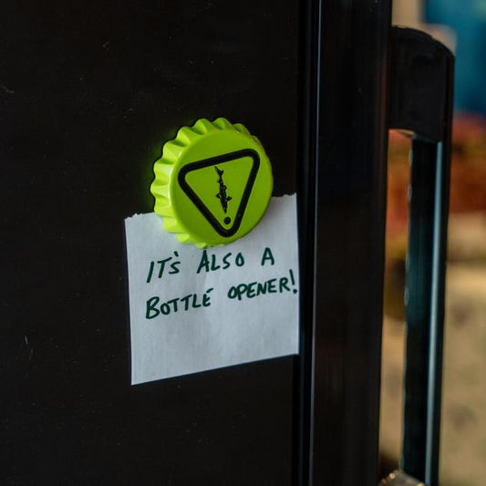The Caution Cap Bottle Opener from Brewery Branding, Inc., shaped like a bottle cap, doubles as a fridge magnet—making it a fun and useful kitchen accessory.