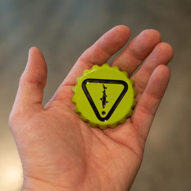 A hand holds the lime green Caution Cap Bottle Opener by Brewery Branding, Inc., with a black warning triangle and lightning bolt, perfect for your kitchen or as a fridge magnet, shown against a blurred background.