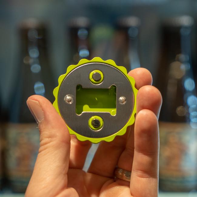 A hand holds a green and silver Brewery Branding, Inc. Caution Cap Bottle Opener, circular in shape with a built-in fridge magnet, with blurred glass bottles in the background.