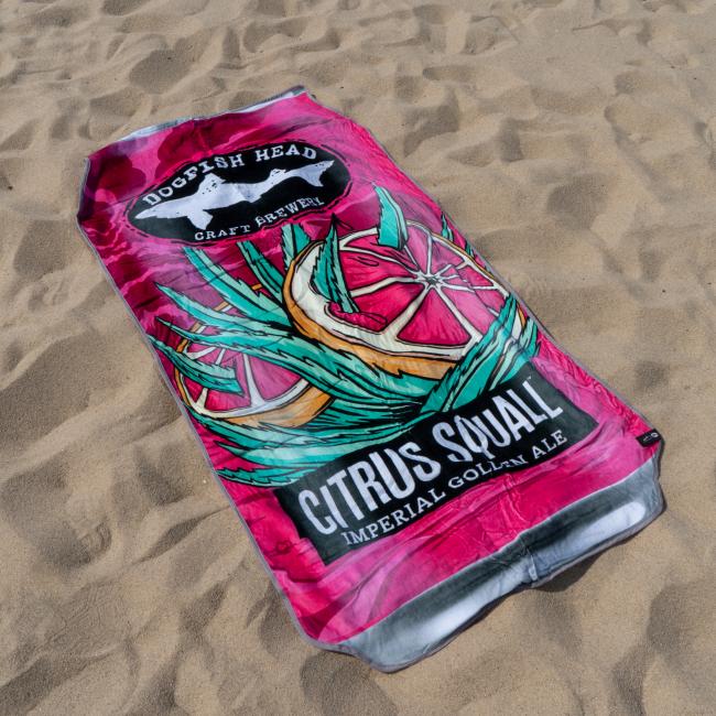 The Citrus Squall Towel by Slowtide, inspired by a Dogfish Head Citrus Squall Imperial Golden Ale can, features citrus slice and agave leaf artwork on soft terry cotton and is shown lying on the sand.