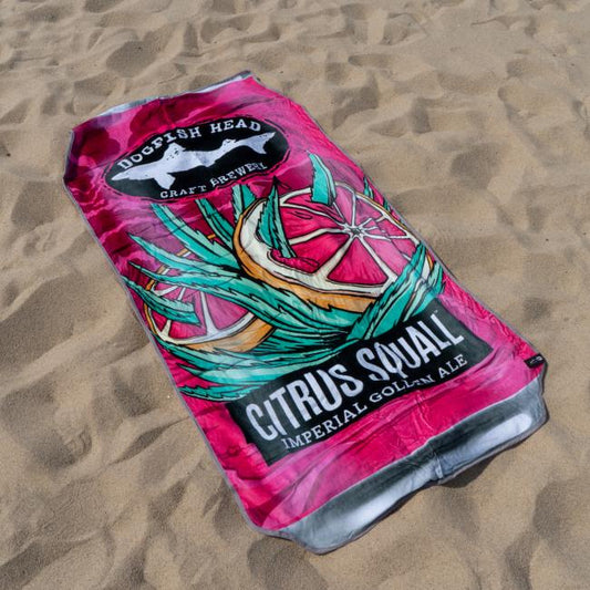 The Citrus Squall Towel by Slowtide, inspired by a Dogfish Head Citrus Squall Imperial Golden Ale can, features citrus slice and agave leaf artwork on soft terry cotton and is shown lying on the sand.
