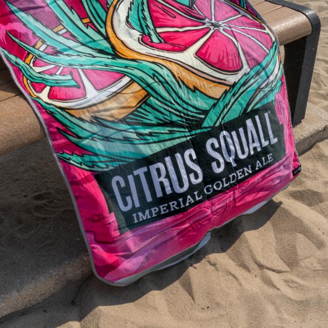 A vibrant Citrus Squall Towel by Slowtide, featuring grapefruit and green leaf designs with "CITRUS SQUALL IMPERIAL GOLDEN ALE" text, is draped over a wooden bench on sandy ground.