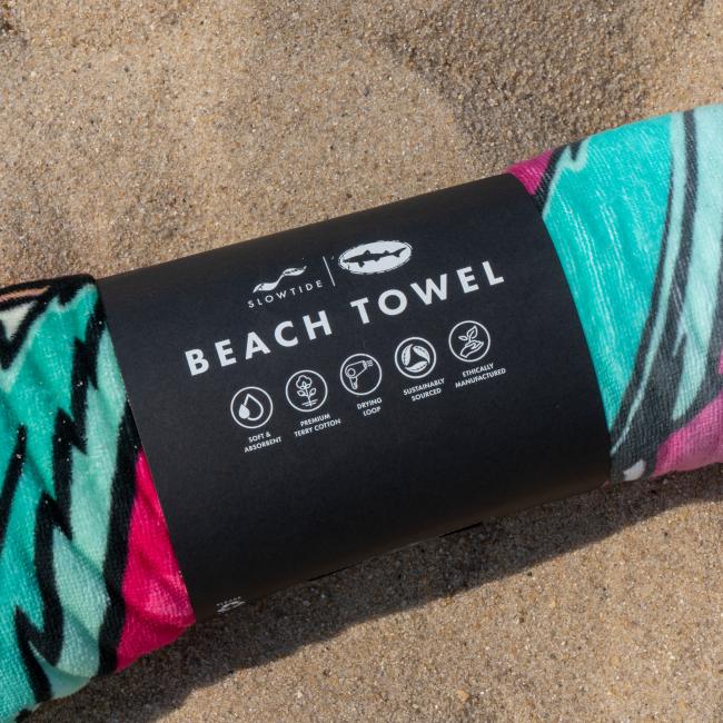 A rolled-up Slowtide Citrus Squall Towel with a colorful pattern rests on sand. Its black label highlights features like soft, absorbent terry cotton, quick drying, and sustainable sourcing.