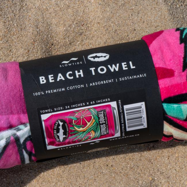 A rolled-up Slowtide Citrus Squall Towel with a black label rests on the sand. The label displays its soft terry cotton material, size, and a small image of the towel’s vibrant, full design.