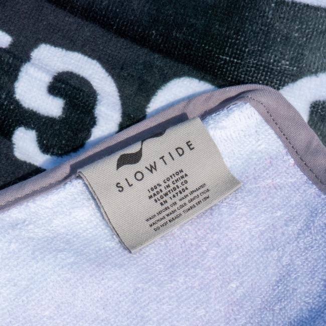 A close-up of the Citrus Squall Towel by Slowtide shows a beige tag with branding and care details. The white terry cotton towel with a gray border is displayed on a dark patterned surface.