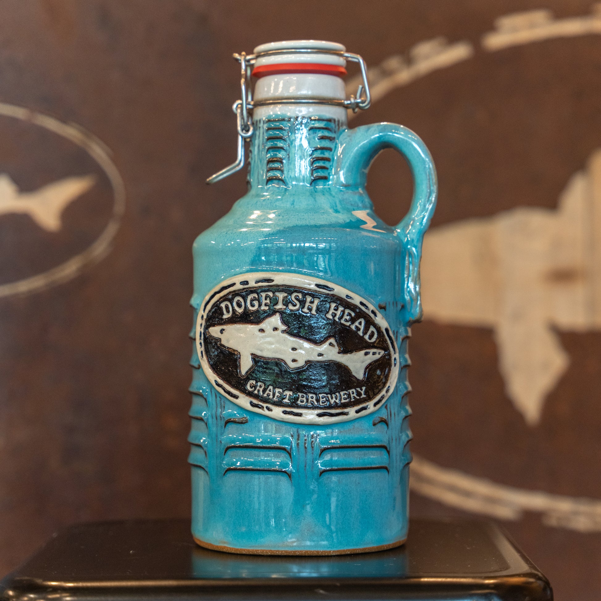A Coastal Blue Ceramic Growler by Romanick Pottery, featuring a handle and wide-mouth hinge top, sits on a dark surface with a brown background displaying a faint fish outline.