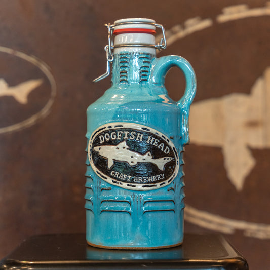 A Coastal Blue Ceramic Growler by Romanick Pottery, featuring a handle and wide-mouth hinge top, sits on a dark surface with a brown background displaying a faint fish outline.