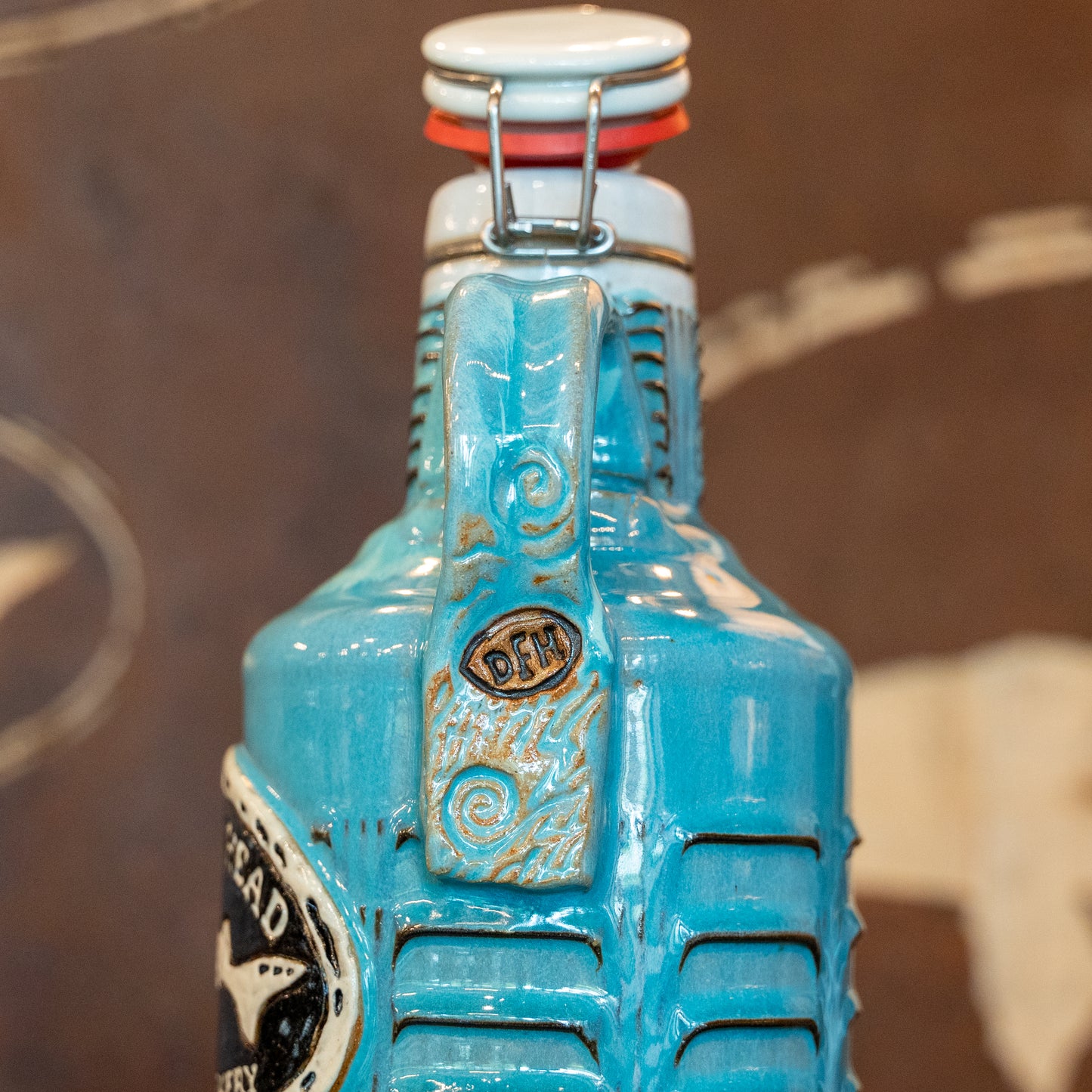 A close-up of the Coastal Blue Ceramic Growler by Romanick Pottery shows its embossed swirls, handle, and "DFH" letters. This handcrafted jug features a wide-mouth hinge top with metal clasp and orange seal. Blurred brown and white background.