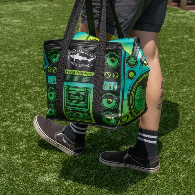 A person in black shorts, striped socks, and slip-on shoes stands on grass holding a Go Match Up Cooler Bag with a retro stereo design. Their tattooed leg is visible next to the colorful insulated soft cooler.