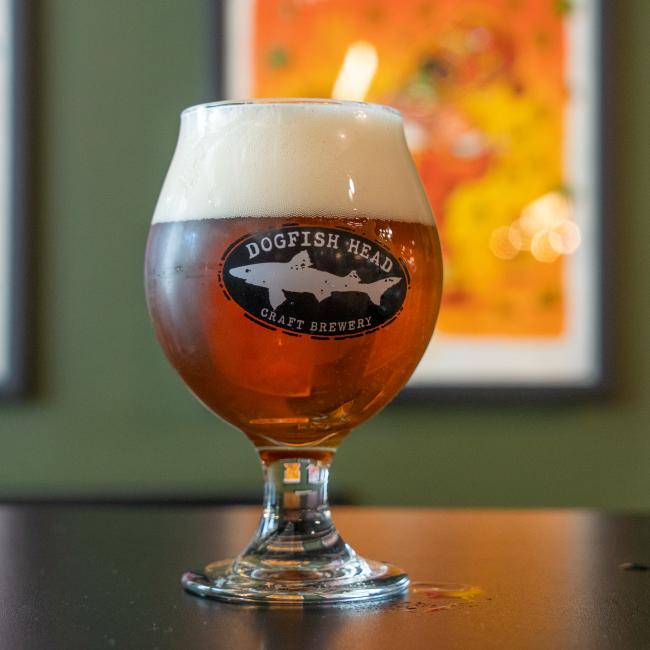The Craft Brewery Occasional Glass by Grandstand Glassware & Apparel, filled with amber beer and a thick foam head, rests on a dark surface with colorful, blurred art in the background.