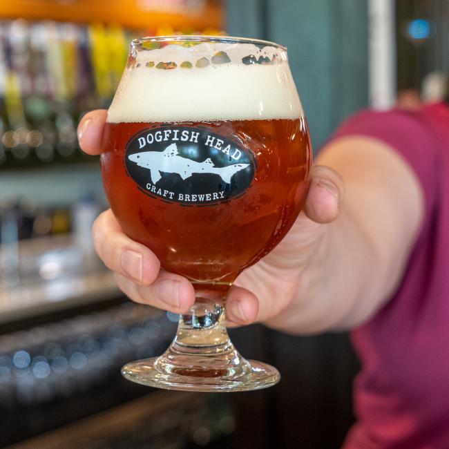 In a bar setting, someone holds a Grandstand Glassware & Apparel Craft Brewery Occasional Glass filled with amber beer and a foamy head.