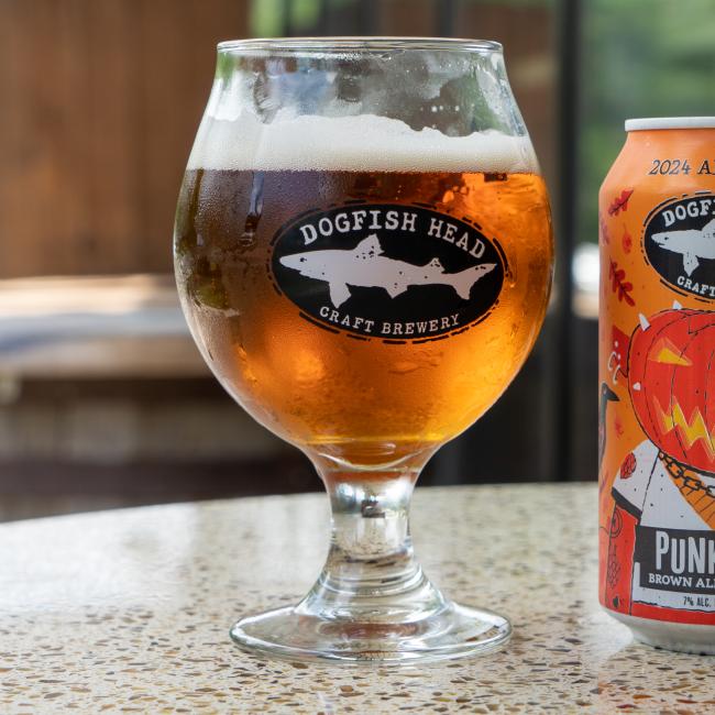 A glass of amber beer with a foamy head, served in the Craft Brewery Occasional Glass from Grandstand Glassware & Apparel, sits on a counter beside a colorful can featuring a pumpkin illustration.