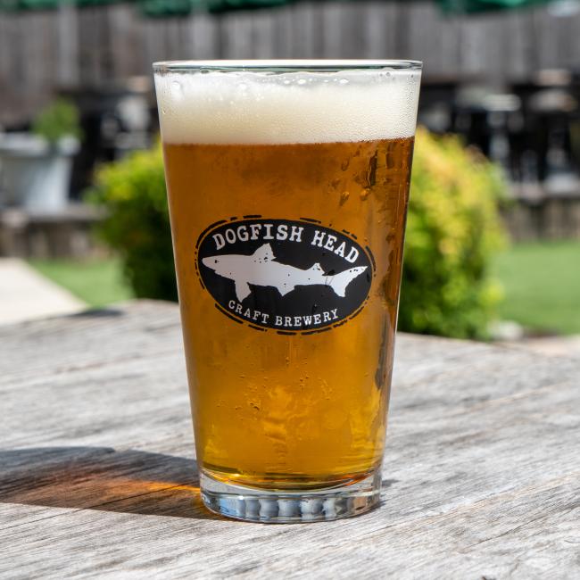 A Merchtank Craft Brewery Pint Glass rests on a wooden outdoor table, with a blurred green background.