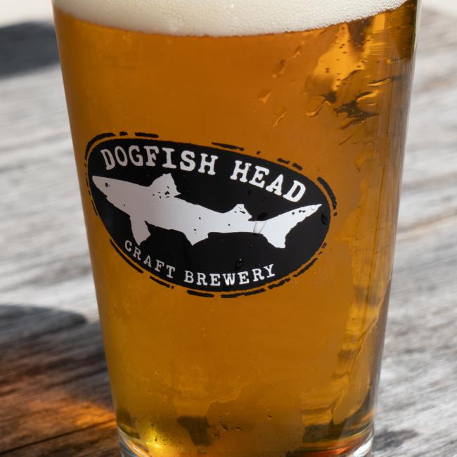 A Merchtank Craft Brewery Pint Glass filled with amber craft beer sits on a wooden surface, featuring the brand's logo and distinctive shark graphic.