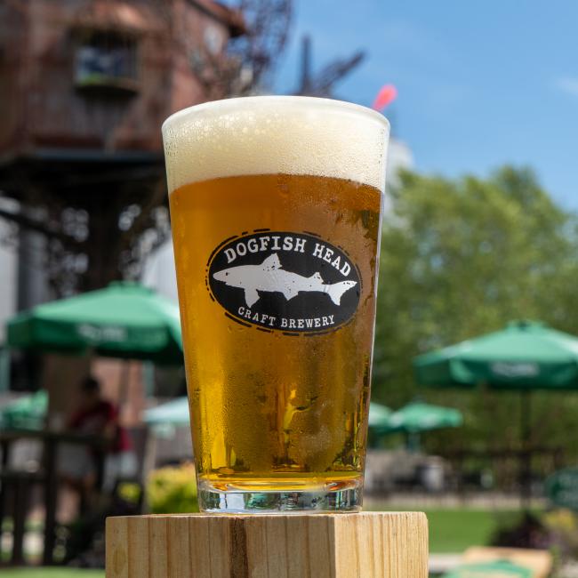 A Craft Brewery Pint Glass from Merchtank, filled with foamy craft beer, rests on a wooden surface outdoors, with green umbrellas and trees in the background.