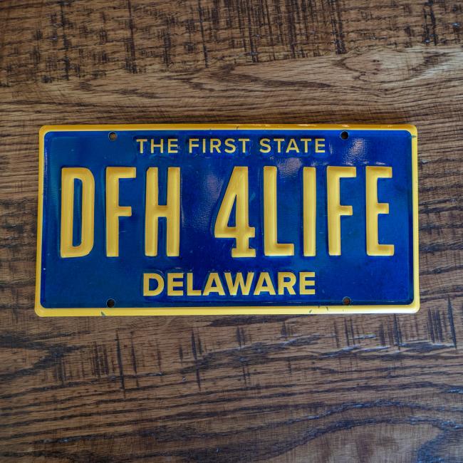Blue license plate tacker with yellow lettering and bordering. License plate "number" is "DFH 4LIFE", state is Delaware and at the top of the tacker "The first state" is written