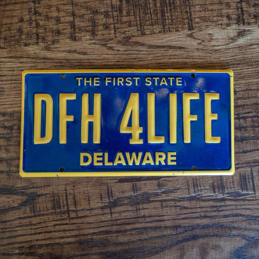 Blue license plate tacker with yellow lettering and bordering. License plate "number" is "DFH 4LIFE", state is Delaware and at the top of the tacker "The first state" is written