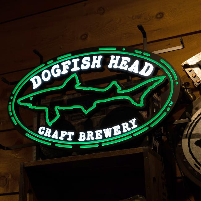 The Merchtank DFH Craft Brewery LED Neon features a bright green shark outline and Dogfish Head Craft Brewery text, glowing vividly on a wooden wall.