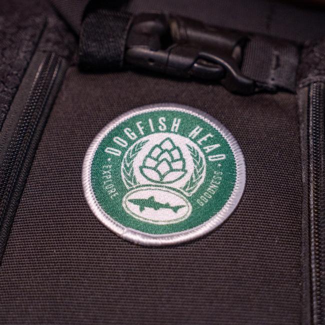 The DFH Explore Goodness Patch by GRAFFITI INC is a round embroidered patch with the Dogfish Head logo, showing a green hop cone and shark silhouette, sewn onto dark fabric near zippers and buckles.