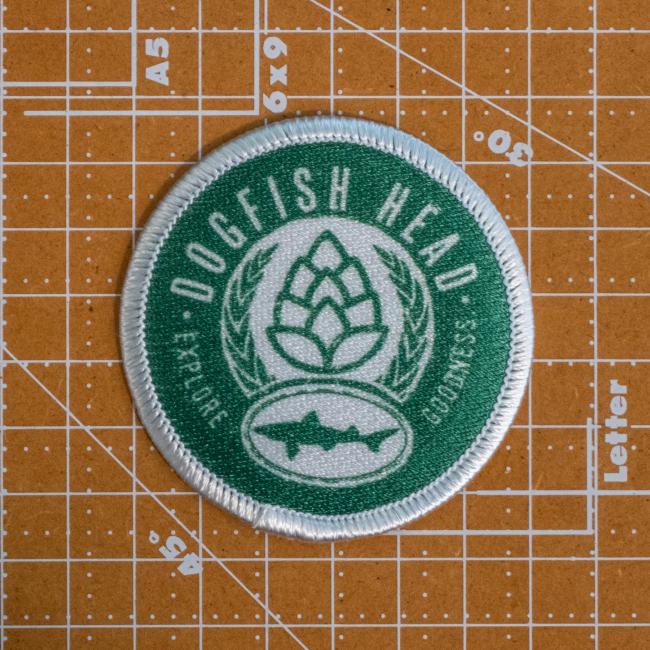 The DFH Explore Goodness Patch by GRAFFITI INC is a round green and white patch featuring Dogfish Head text, a hop and fish graphic—ideal for those aiming to explore goodness in every sip.
