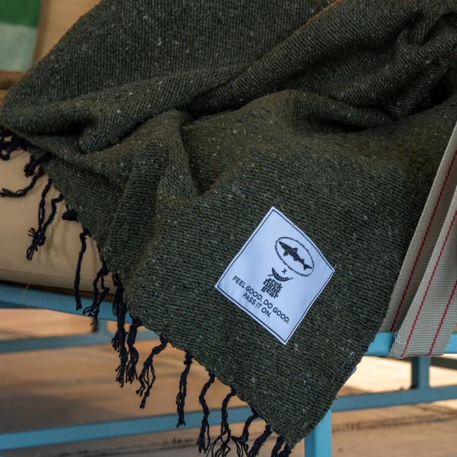 A green INN Blanket by Trek Light Gear with black fringe drapes over a tan cushion. A white patch on the upcycled blanket reads “FEEL GOOD. DO GOOD. PASS IT ON.” with a fish image above the text.