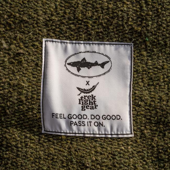 A white fabric label sewn onto textured green material shows a fish logo above “Trek Light Gear” and “Feel good. Do good. Pass it on.” in black, accenting the handmade INN Blanket crafted from recycled materials.