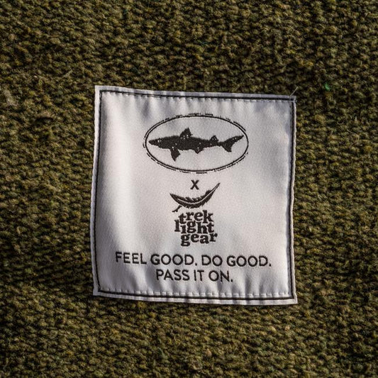 A white fabric label sewn onto textured green material shows a fish logo above “Trek Light Gear” and “Feel good. Do good. Pass it on.” in black, accenting the handmade INN Blanket crafted from recycled materials.