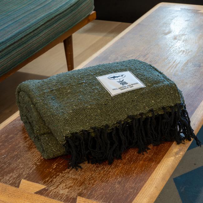 The INN Blanket by Trek Light Gear, in dark green with black fringe, is neatly folded on a wooden table. A white patch label with a logo and text is visible, and a green upholstered bench appears in the background.