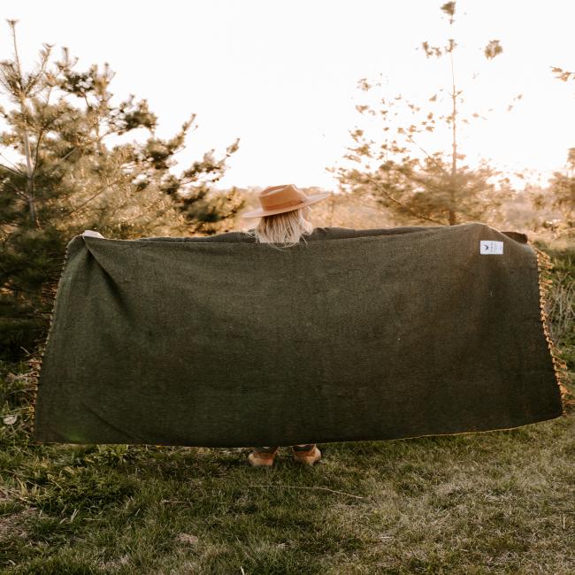 A person outdoors on grass holds up a large dark green Trek Light Gear INN Blanket, surrounded by trees and greenery in soft sunlight.