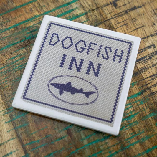 Coaster with a sort of needlepoint pattern and a white border. The coaster has writing "Dogfish Inn" in blue and the shark and shield logo below it.
