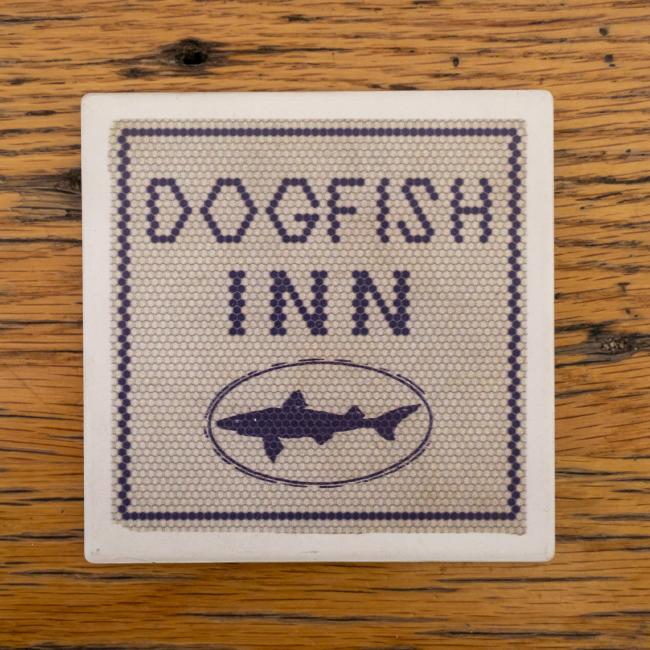 Coaster with a sort of needlepoint pattern and a white border. The coaster has writing "Dogfish Inn" in blue and the shark and shield logo below it.