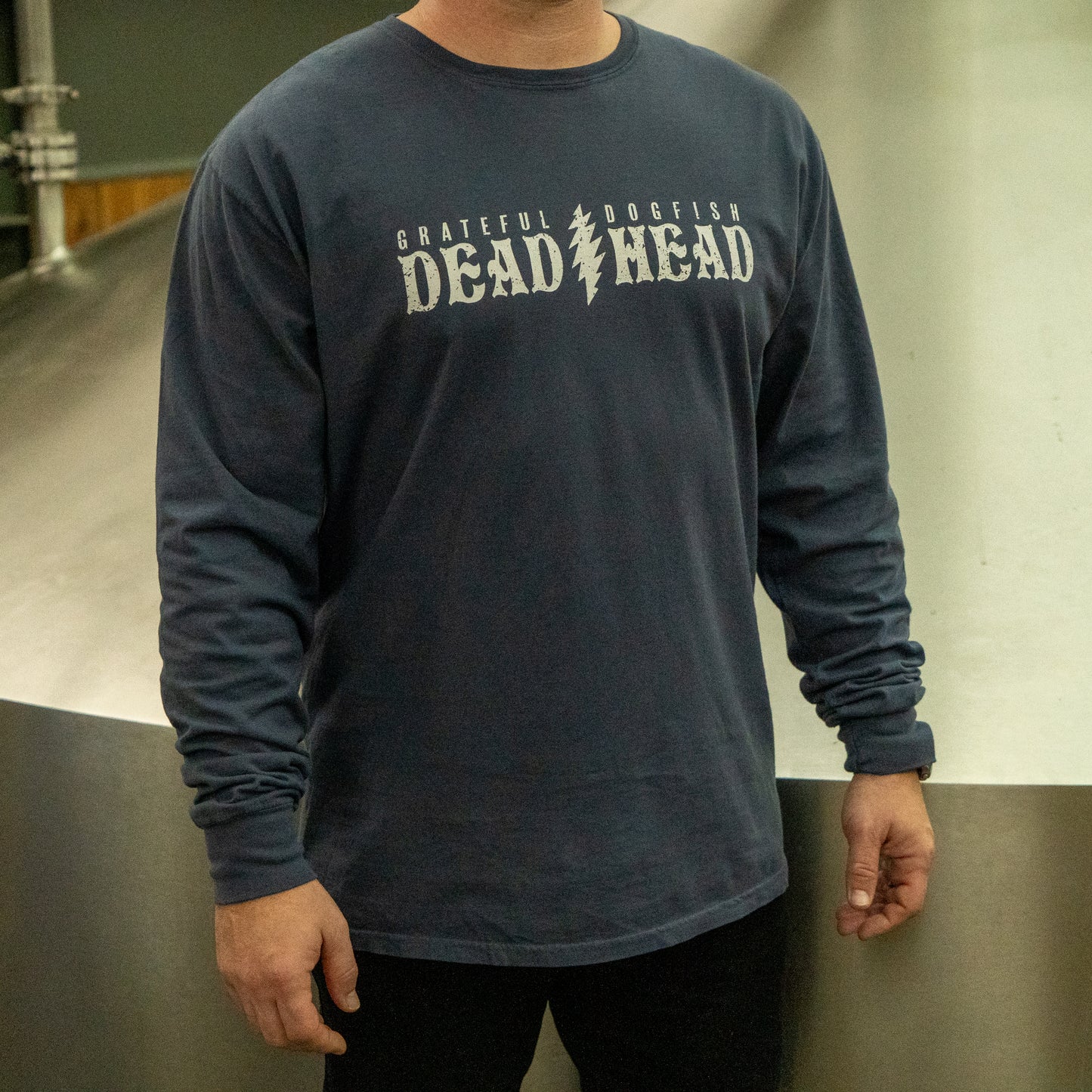 Someone wearing a Dogfish Head x Grateful Dead Anchor Slate Long Sleeve Tee by GO WEST T-SHIRT COMPANY stands indoors beside metal tanks.