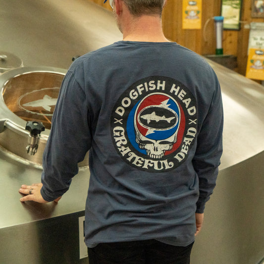 Wearing the Dogfish Head x Grateful Dead Anchor Slate Long Sleeve Tee by GO WEST T-SHIRT COMPANY, a person stands with their back to the camera beside a large metal brewing tank in a brewery.