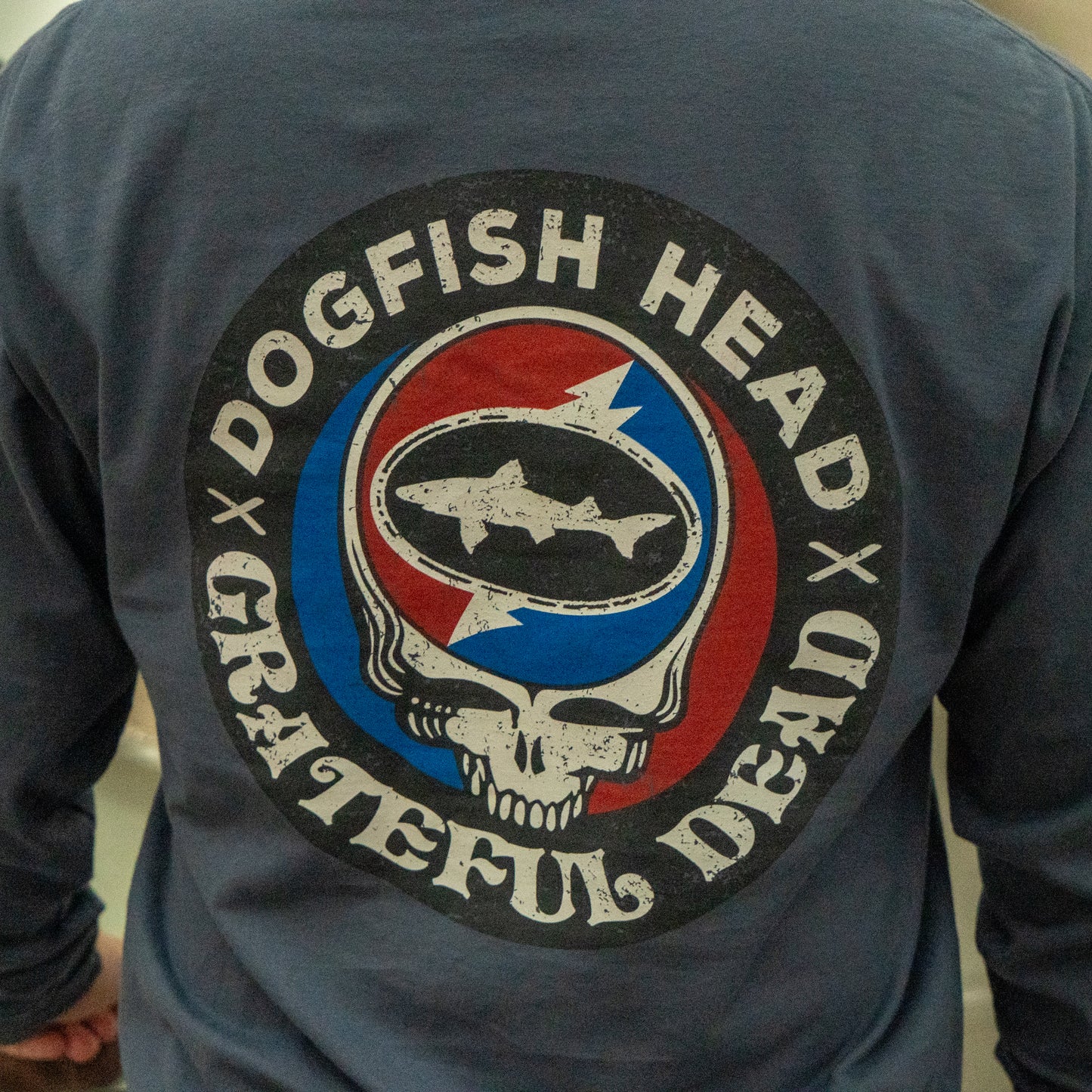 A person wears the Dogfish Head x Grateful Dead Anchor Slate Long Sleeve Tee by GO WEST T-SHIRT COMPANY, featuring a circular shark and skull logo in red, white, and blue with "Dogfish Head Grateful Dead" around the design.