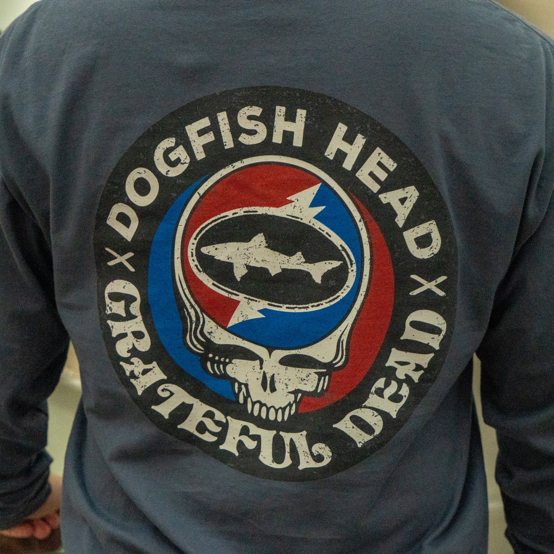 A person wears the Dogfish Head x Grateful Dead Anchor Slate Long Sleeve Tee by GO WEST T-SHIRT COMPANY, featuring a circular shark and skull logo in red, white, and blue with "Dogfish Head Grateful Dead" around the design.