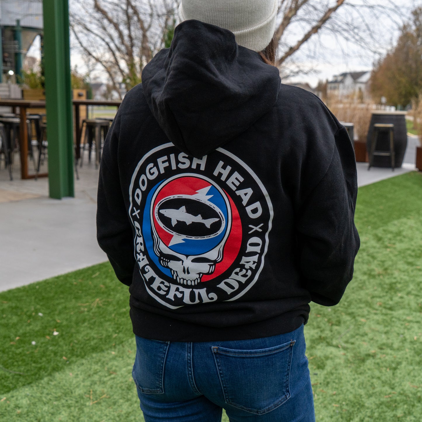 Someone stands on the grass in a GO WEST T-SHIRT COMPANY Dogfish Head x Grateful Dead Black Hoodie, featuring a fish and skull graphic inside a red, white, and blue circle on the back.