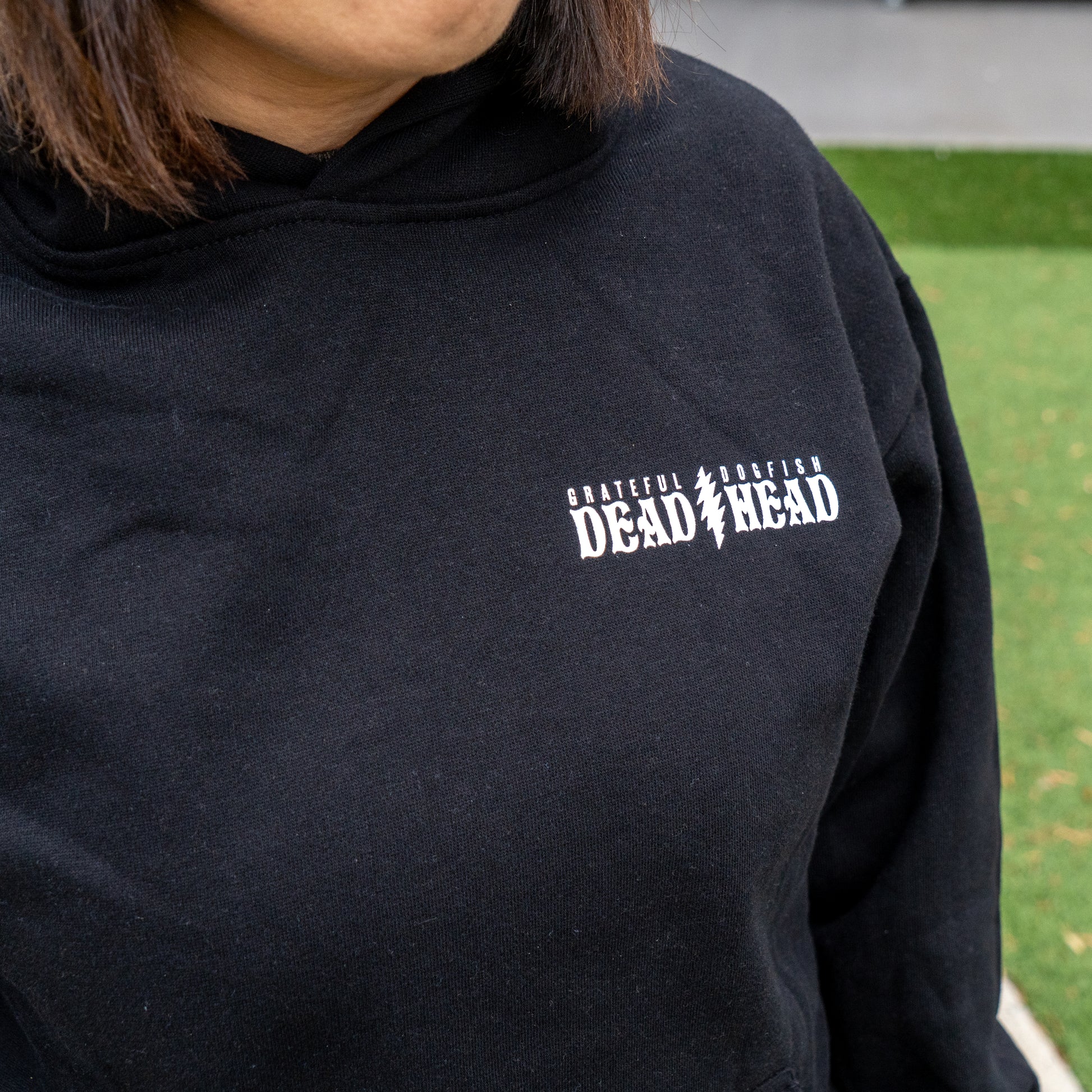 A person stands on grass wearing a GO WEST T-SHIRT COMPANY Dogfish Head x Grateful Dead Black Hoodie with "GRATEFUL MORE FISH DEAD HEAD" in white on the chest; their face is partially out of frame.