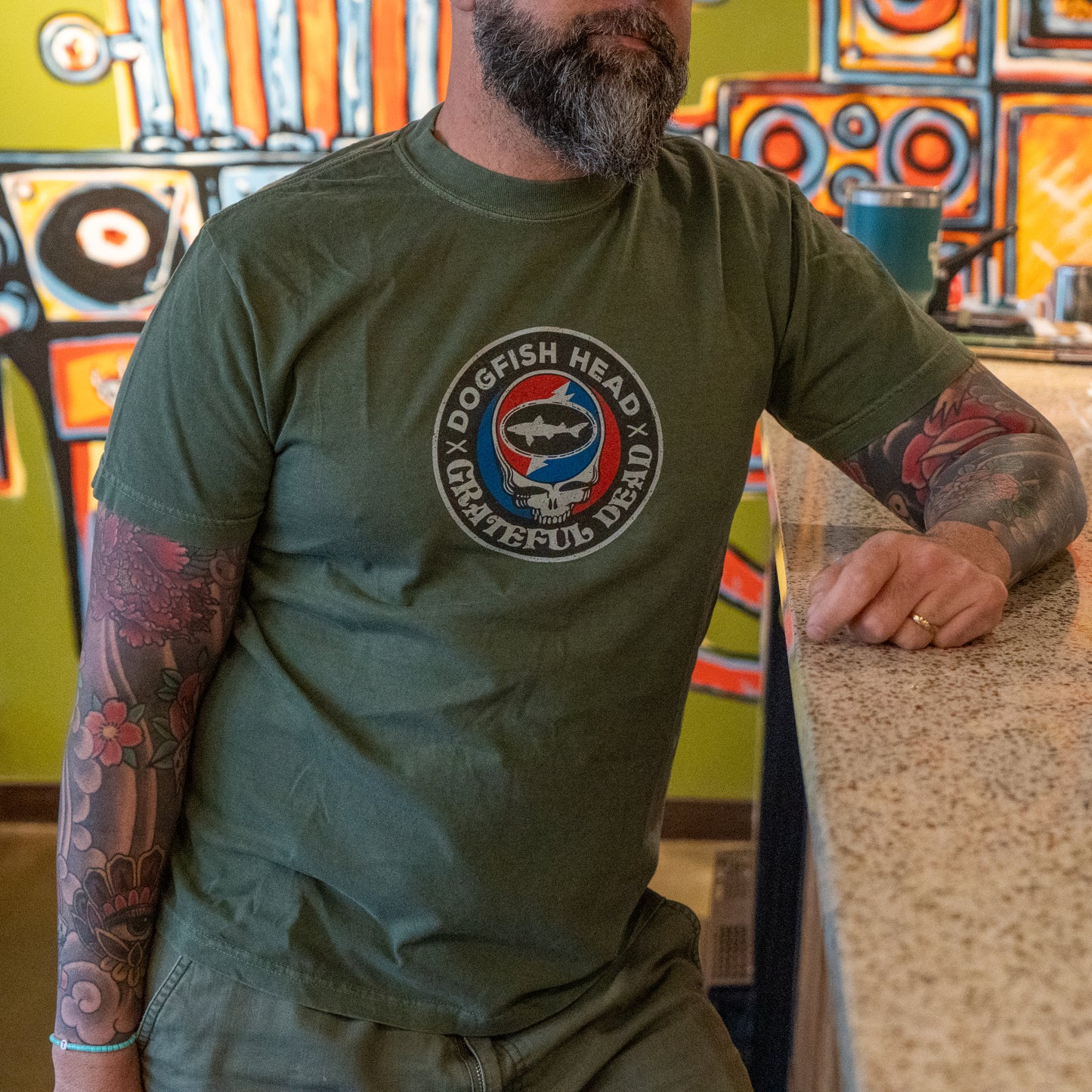 A bearded man with tattooed arms leans on a countertop, wearing a GO WEST T-SHIRT COMPANY Dogfish Head x Grateful Dead Green Logo Tee. Colorful abstract artwork is visible on the wall behind him.