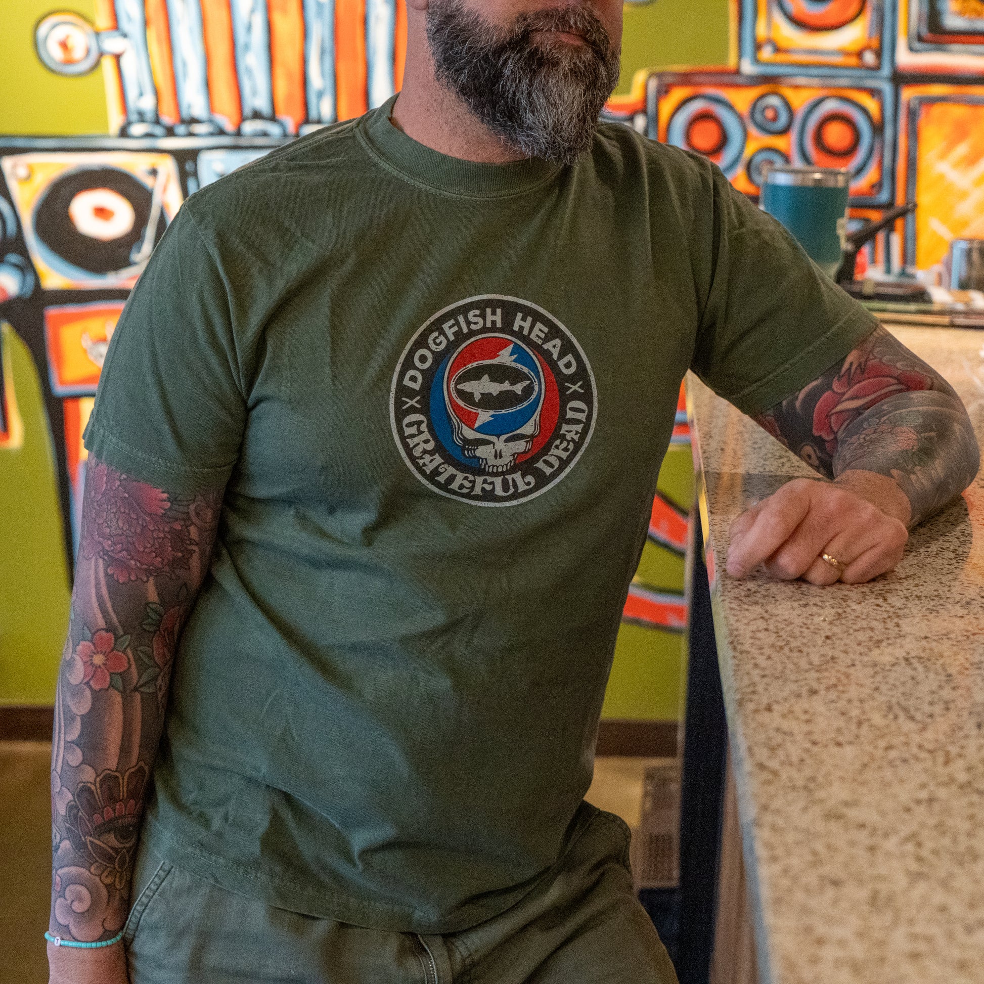 A bearded man with tattooed arms leans on a countertop, wearing a GO WEST T-SHIRT COMPANY Dogfish Head x Grateful Dead Green Logo Tee. Colorful abstract artwork is visible on the wall behind him.