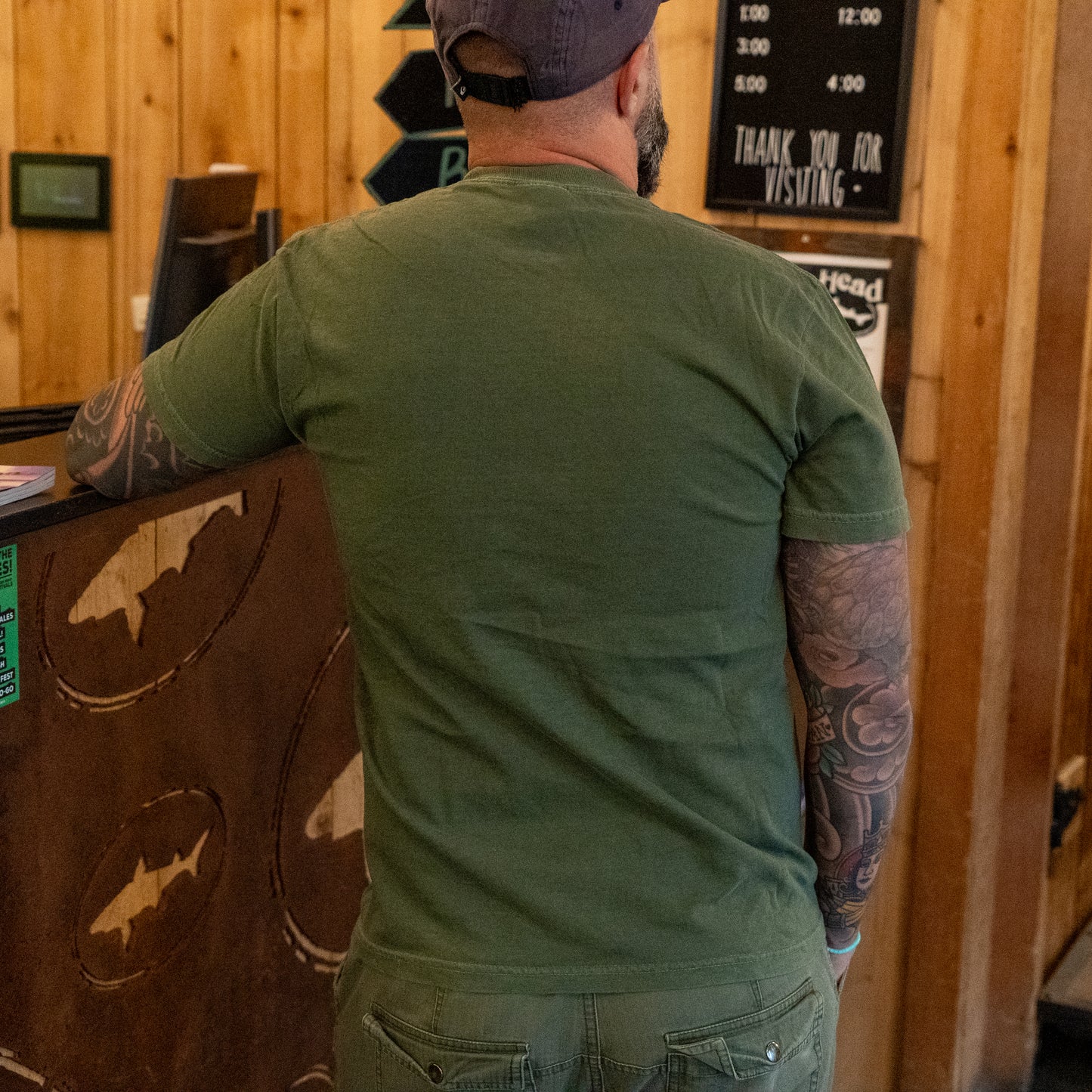 Wearing a Dogfish Head x Grateful Dead Green Logo Tee by GO WEST T-SHIRT COMPANY, a tattooed individual stands with their back to the camera at a rustic wooden counter, also sporting a green cap.
