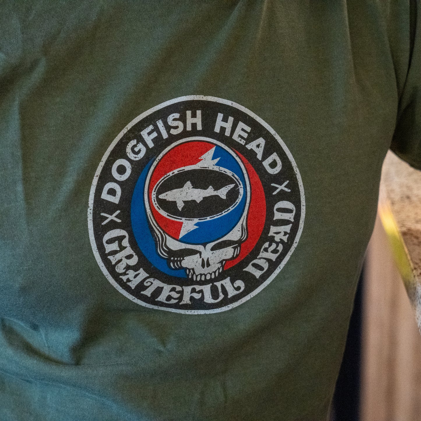 The Dogfish Head x Grateful Dead Green Logo Tee by GO WEST T-SHIRT COMPANY is a soft ring-spun cotton t-shirt featuring a circular logo with both brands, a stylized skull, fish silhouette, and red/blue lightning bolt background.