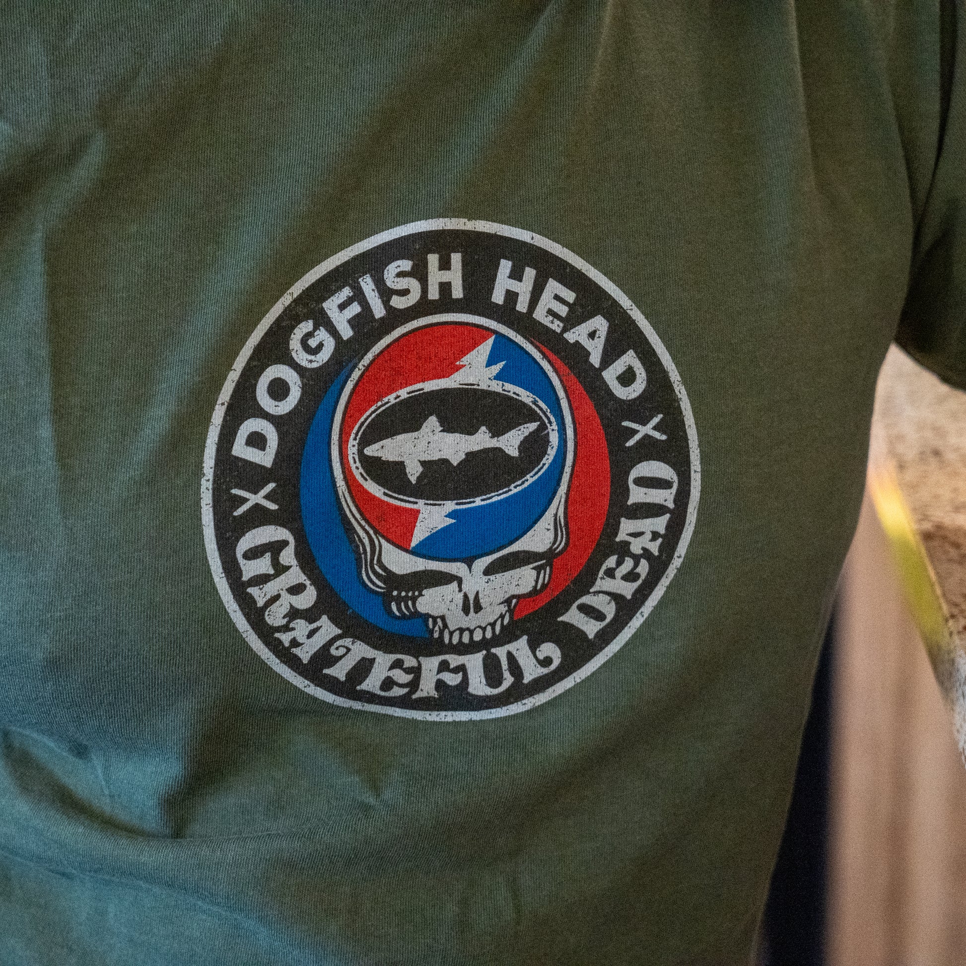 The Dogfish Head x Grateful Dead Green Logo Tee by GO WEST T-SHIRT COMPANY is a soft ring-spun cotton t-shirt featuring a circular logo with both brands, a stylized skull, fish silhouette, and red/blue lightning bolt background.